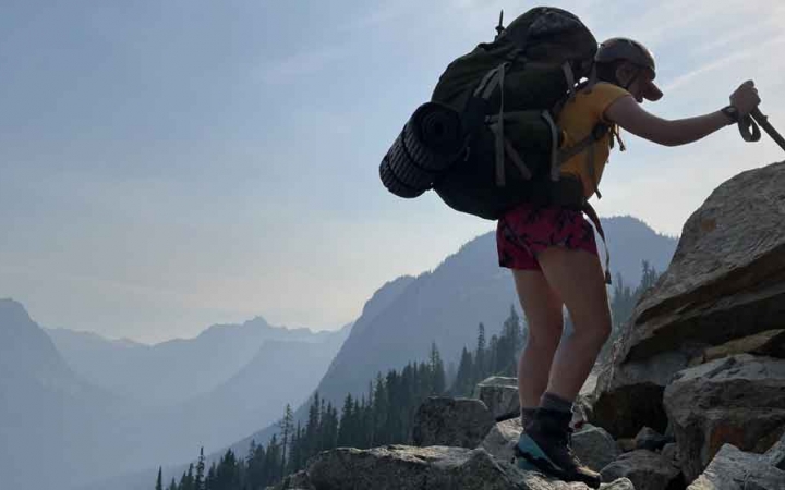 a person carrying a backpack braces themself using a trekking pole. There are mountains in the background. 